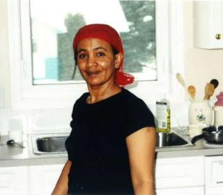 A woman wearing a headscarf stands in her kitchen smiling at the camera.