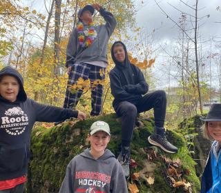 Five boys in hoodies hang out around a large rock in a forest