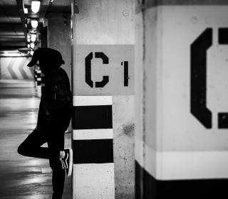 In this black and white photograph, a young man in a hoodie looks pensive while he leans against the wall in a subway station. 