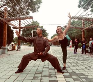 The author, a young White woman, balances in a ballet pose holding the shoulder of an elderly Tai Chi master in the streets of Jiuquan, China while amused people look on.