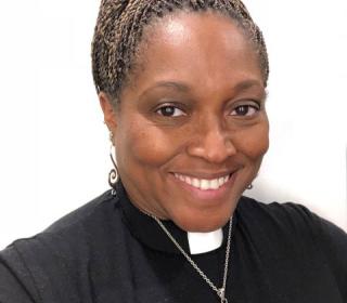 A portrait of Rev. Dr. Karen Georgia Thompson, a Black woman with beautiful braids atop her head in a bun, wearing a clergy collar and a metal owl pendant.