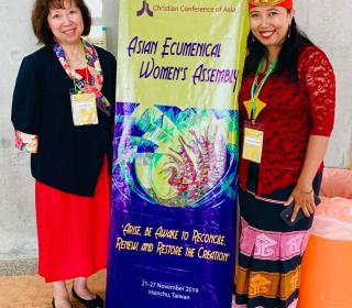 Author Kim Uyede-Kai from the United Church stands with Rev. Ester Damaris Wolla Wunga from Indonesia stand with an assembly banner. Both women are of Asian background, but the "traditional dresses" they are wearing reflect their different cultures.