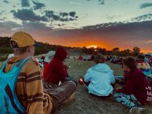 A group of young people sit watching a brilliant, colourful sunset.