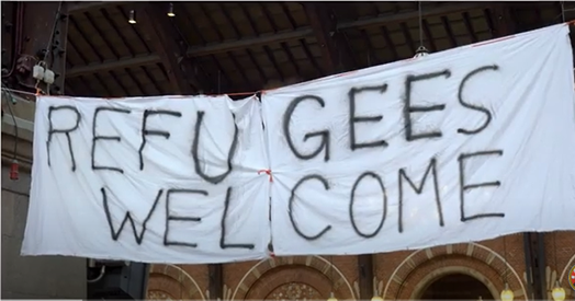 A banner held up by 2 people reads Welcome Refugees