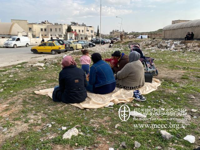 A group of women sit on a blanket in the middle of rubble
