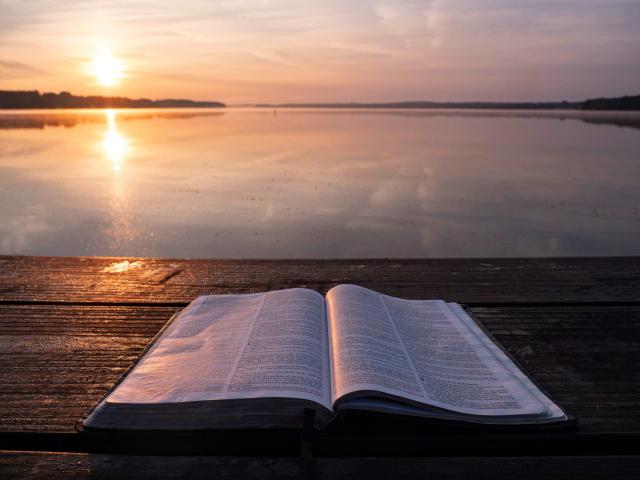 Open Bible lies on dock. The sun sets over a lake in the background.