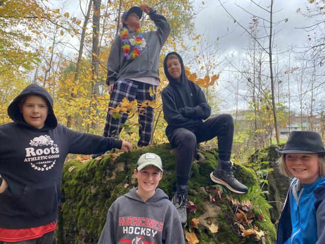 Five boys in hoodies hang out around a large rock in a forest
