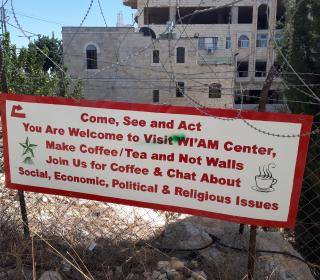   A welcome sign for Wi'am Center on a barbed-wire fence in Palestine, with the message, "Make Coffee/Tea and Not Walls."