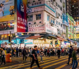 A busy night street scene in Hong Kong, with large groups of people crossing an intersection, backed by well-lit buildings covered with advertisements and signs.