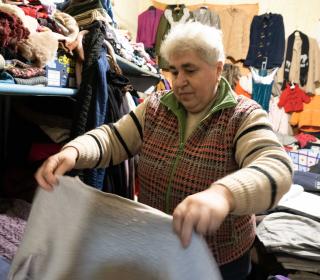 A woman with short white hair holds up clothes while folding them, in a store room of supplies for Ukraine refugees.