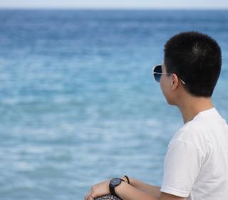 A man in a white shirt and sunglasses sits on a seawall and looks out to sea on a bright, sunny day against a deep blue sky.
