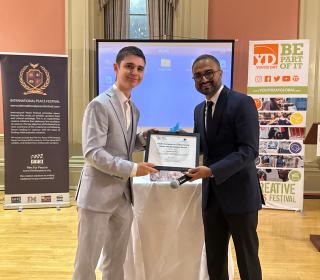 A young man in a suit receives a plaque from another man in a suit at an awards ceremony.