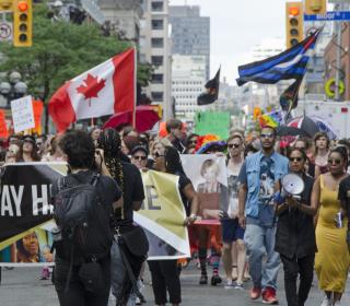 Black Lives Matter supporters march at Toronto Pride 2016, beneath a large Canadian flag and flags representing the Trans movement.