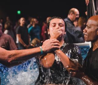 A diverse group of leaders pull a young person holding his nose out of the baptism waters. A number of people in the background await their turn to be baptized.