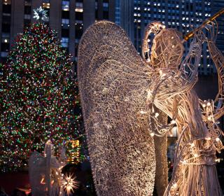 A sculpture of an angel blowing a horn, standing in front of a Christmas tree with multi-coloured lights.