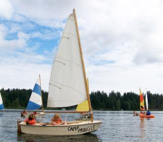 A small sailboat being piloted by two children in lifejackets in a lovely lake surrounded by forest. In the background are a number of similar boats piloted by camp kids.