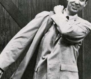 A black and white photograph of Fred Rogers taken in the late 1960s. He is a White man with dark hair and an inviting smile. He is shown wearing a light coloured suit jacket while pulling a bag over his shoulder.