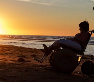 A person in a beach wheelchair crosses the sand while silhouetted by the brillant sunset.