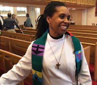 Rev. B. Maya Douglas, a Black woman, stands smiling among the pews of her church, wearing an alb and colourful embroidered stole.