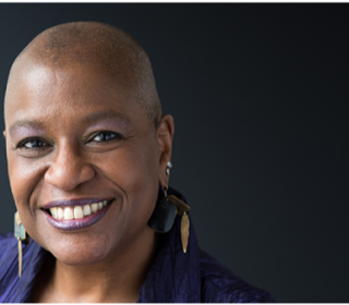 Dr. Velda Love of the United Church of Christ, a Black woman in an indigo wrap, with very closely cropped hair, a brilliant smile, and long earrings, looks directly at the camera.