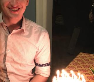 A young white man standing in a door frame, illuminated by the candles on the cake for his birthday.