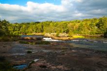 A view at a wide river, with a small falls in the middle. The foreground is smooth round rocks shaped by the water. The background are the trees of a forest on the distant bank, brilliantly highlighted by the sunlight.
