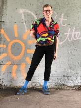 A young White man with short light brown hair, in a bright multi-colour shirt, black pants, and tall shocking blue socks, poses energetically before a wall with graffiti on it.