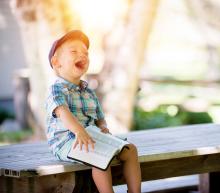 A young white boy with a cap and a Bible in his lap laughs out loud while seated at an empty picnic table.