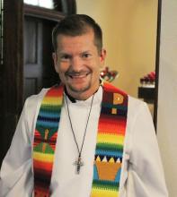 A portrait of the Rev. Jeff Rock of Metropolitan Community Church, a young White man with short light hair. Wearing a white alb and rainbow stole.