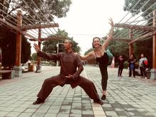 The author, a young White woman, balances in a ballet pose holding the shoulder of an elderly Tai Chi master in the streets of Jiuquan, China while amused people look on.