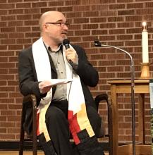 Moderator Richard Bott, on stage, seated in a chair and gesturing to his left while speaking. He wears a colourful stole