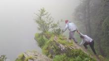 Two people work collaboratively to climb over a rock on a foggy forest mountain.