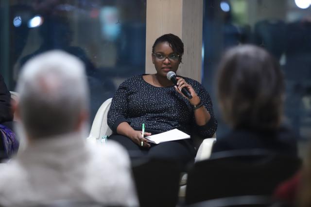 Jane Thirikwa, a Black woman with glasses and dressed in black, speaks at the event, sitting while holding a microphone and a pad of paper. She is foreshadowed by members of the audience.
