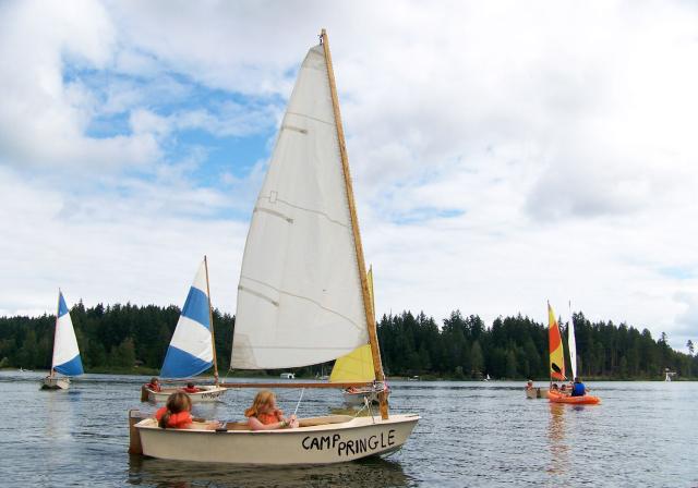 Sailing at Camp Pringle, Vancouver Island A small sailboat being piloted by two children in lifejackets in a lovely lake surrounded by forest. In the background are a number of similar boats piloted by camp kids.