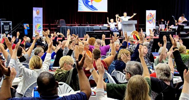 Closing worship at General Council 43, July 27, 2018 A group of people sitting at tables holding their arms in the air.