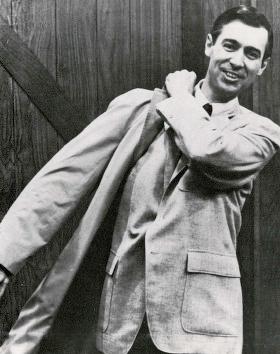 Fred Rogers, late 1960s A black and white photograph of Fred Rogers taken in the late 1960s. He is a White man with dark hair and an inviting smile. He is shown wearing a light coloured suit jacket while pulling a bag over his shoulder.