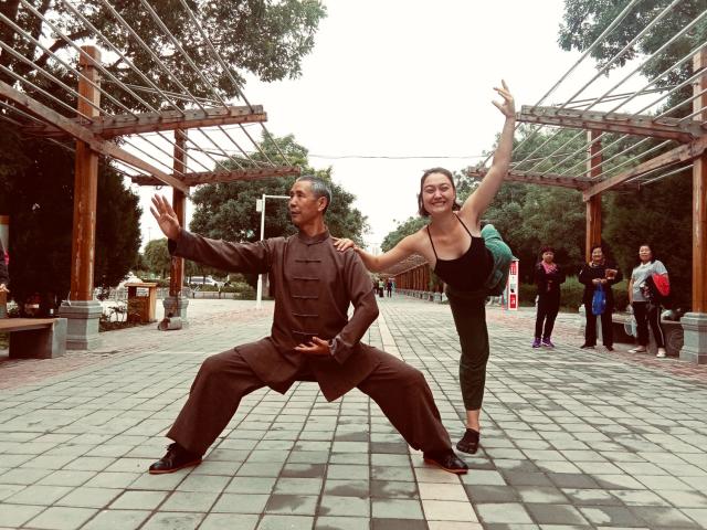 The author (right) shares her ballet skills with a Tai Chi master in Jiuquan, China. The author, a young White woman, balances in a ballet pose holding the shoulder of an elderly Tai Chi master in the streets of Jiuquan, China while amused people look on.