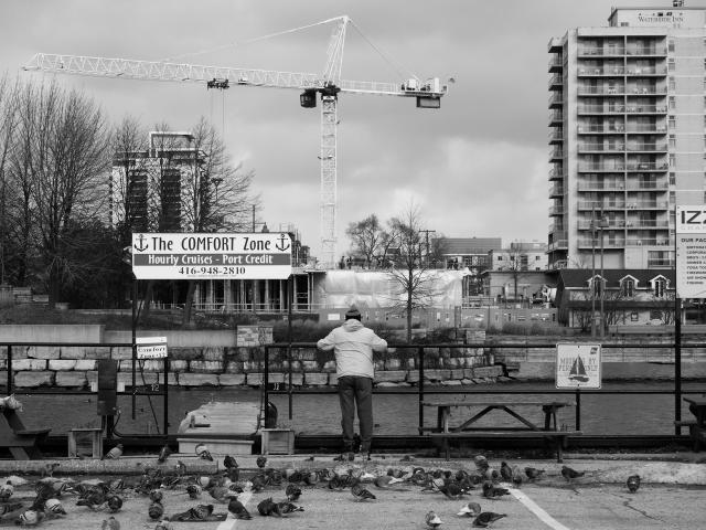 In this black and white photograph, a man is seen from behind as he leans against a railing overlooking a small but busy marina. On the other side of the water is a tall hotel, and next to it, a tall crane where a new development is in the works.