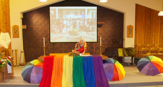 The altar of St. Peter's United Church in Sudbury, ON decorated for a Pride celebration. A minister stands in front of a church altar decorated for a Pride celebration with rainbows.